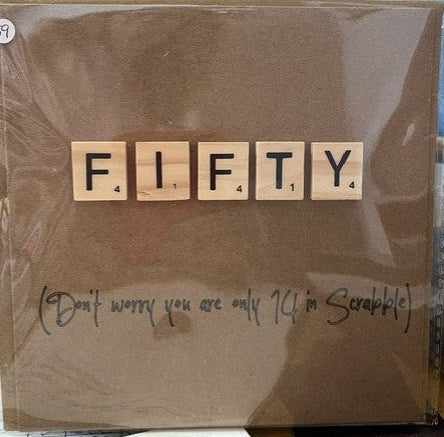 Card with 'FIFTY' spelled out using wooden tiles, with a humorous note underneath, on a table.