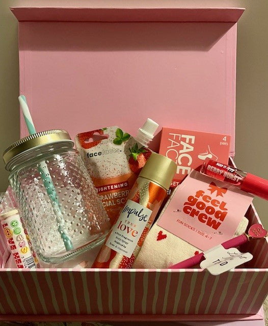 Gift basket with various items including a glass drinking cup, face wash, body spray, socks, pen, lip gloss and candy in a pink & white box.