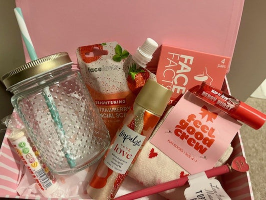 Gift basket with various items including a glass drinking cup, face wash, body spray, socks, pen, lip gloss and candy in a pink & white box.