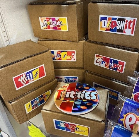 Stack of cardboard boxes containing mugs with the various candy brand words on. Ceramic coaster with Titties on top of Babe mug box.